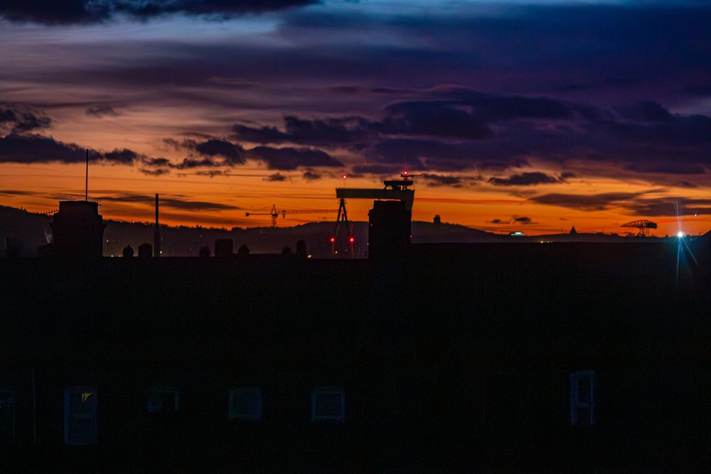 A reddish-orange glow on the pre-dawn horizon, with silhouettes of the Belfast skyline in the foreground