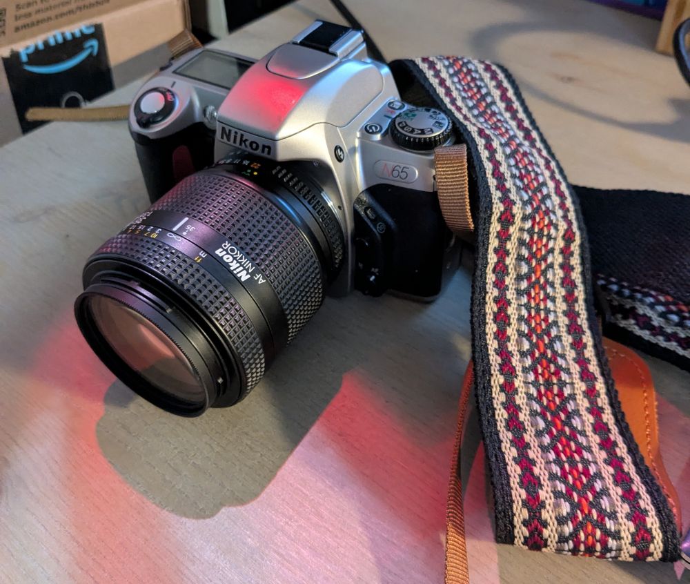 A tan and black N65 Nikon camera with a telephoto lens and patterned camera strap