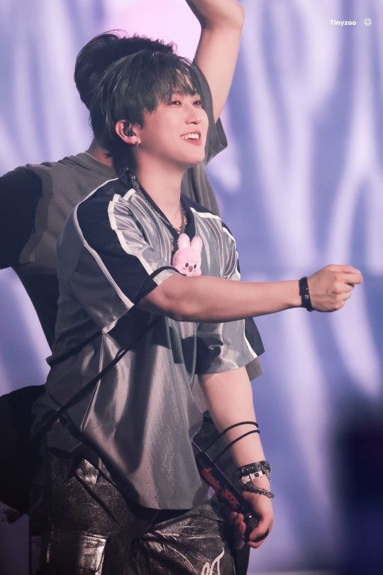 Changbin is dressed in a silver shirt with a black stripe and white stripes on the sleeves, gray jeans, and Dwaekki is hanging from his shirt. He is performing the song 'I Like It.'