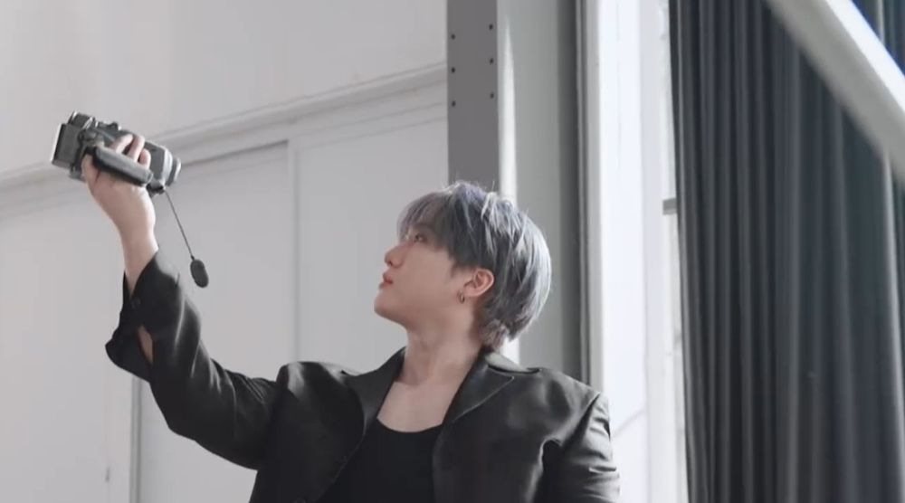 Changbin is dressed in a black blazer and a black t-shirt. He has a small hoop earring in his left ear. His hair is purple. And he is looking at the video camera in his hand, as if he is filming himself