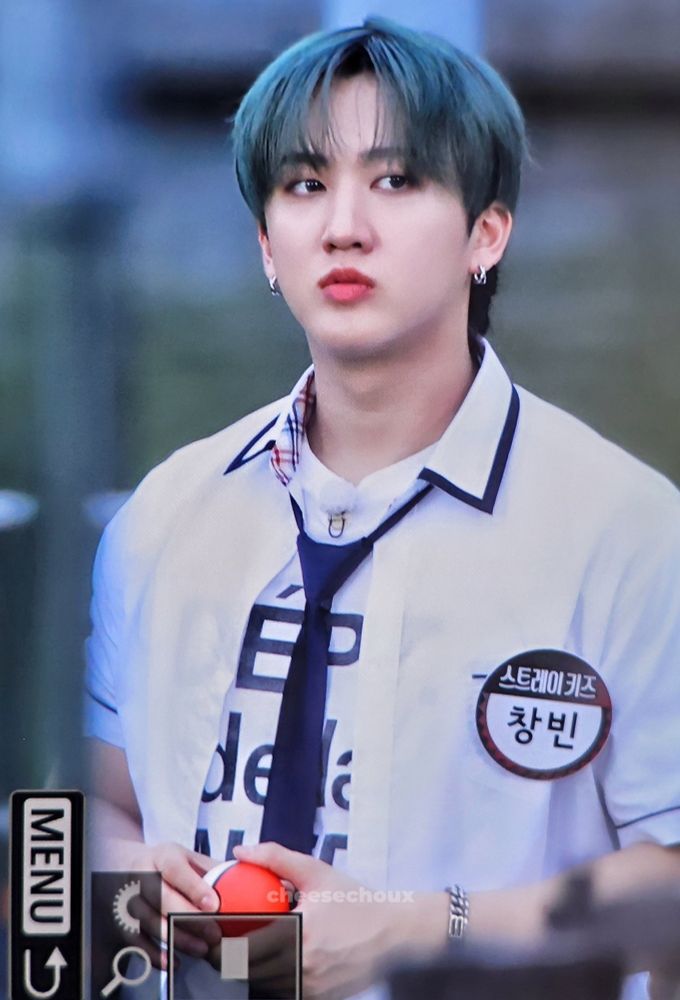 Changbin is wearing a white shirt with a black lettering, a schoolboy-style button-down shirt with a collar, and a navy blue tie. His hair is green. He's holding a pokebola