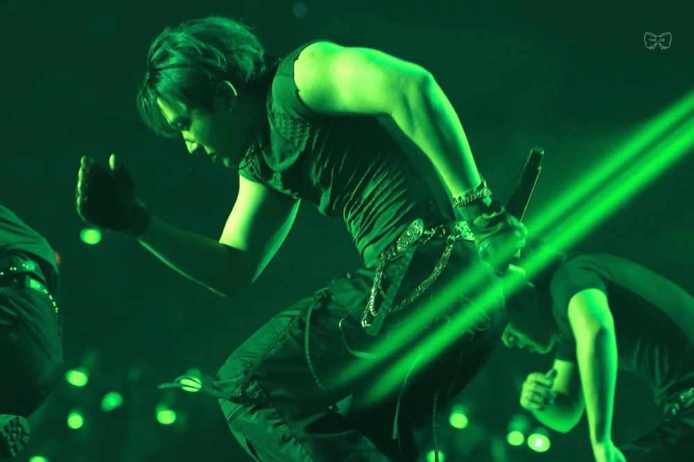 Changbin is wearing a black sleeveless compression shirt, black pants, and a black belt with metallic dots. He is performing his solo. The image is greenish due to the stage lights.