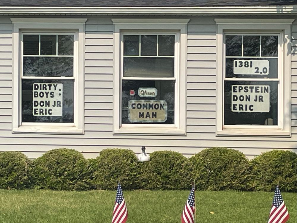 House with gray clapboard siding with miniature USA flags in front of a lawn and shrubs 

3 windows with signs that read

DIRTY BOYS :
DON JR
ERIC
QANON 
COMMON
MAN
1381
2.0
EPSTEIN
DON JR
ERIC