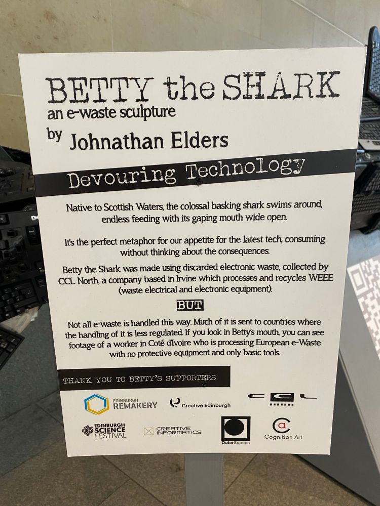 A sign reading “BETTY the SHARK
an e-waste sculpture by Johnathan Elders
Devouring
Technology
Native to Scottish Waters, the colossal basking shark swims around, endless feeding with its gaping mouth wide open.
Its the perfect metaphor for our appetite for the latest tech, consuming without thinking about the consequences.
Betty the Shark was made using discarded electronic waste, collected by CCL North, a company based in Irvine which processes and recycles WEEE (waste electrical and electronic equipment).
BUT
Not all e-waste is handled this way. Much of it is sent to countries where the handling of it is less regulated If you look in Betty's mouth, you can see footage of a worker in Coté dilvoire who is processing European e-Waste with no protective equipment and only basic tools.”