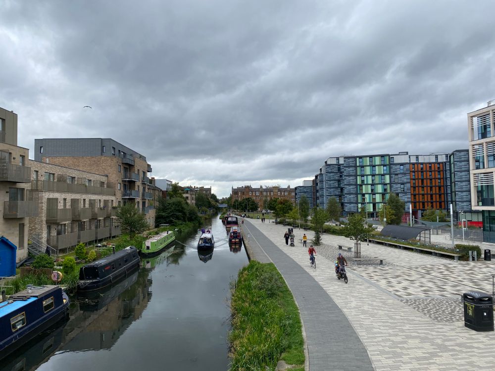 More canal boats as we look west. Modern apartment blocks line both sides of the river. The new school and modernised towpath lie on the right, with cyclists and walkers frequenting the path.