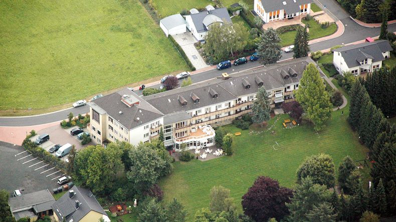 Schräg von oben aus Nordwesten das langgestreckte Anwesen von Haus Höhenblick. Im Norden schließt sich ein Parkplatz an. Am Anwesen führt im Osten eine Straße entlang. Im Westen befinden sich ein Rasen sowie Bäume und Gebüsch.