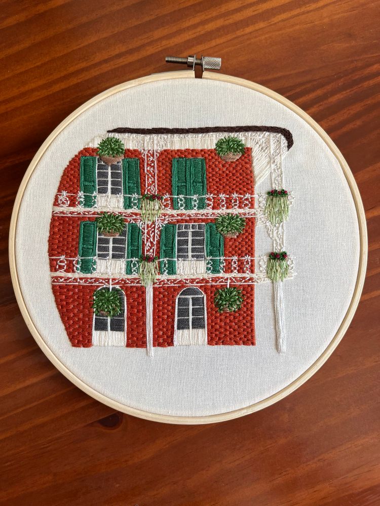 Embroidery art of a New Orleans building, featuring burnt orange brick, green window shutters, and several decorative planters.
