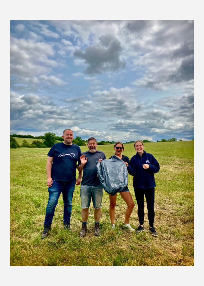 4 people stand in a field with cloudy skies overhead. They are all looking at the camera and 1 of them is holding a black bin liner.