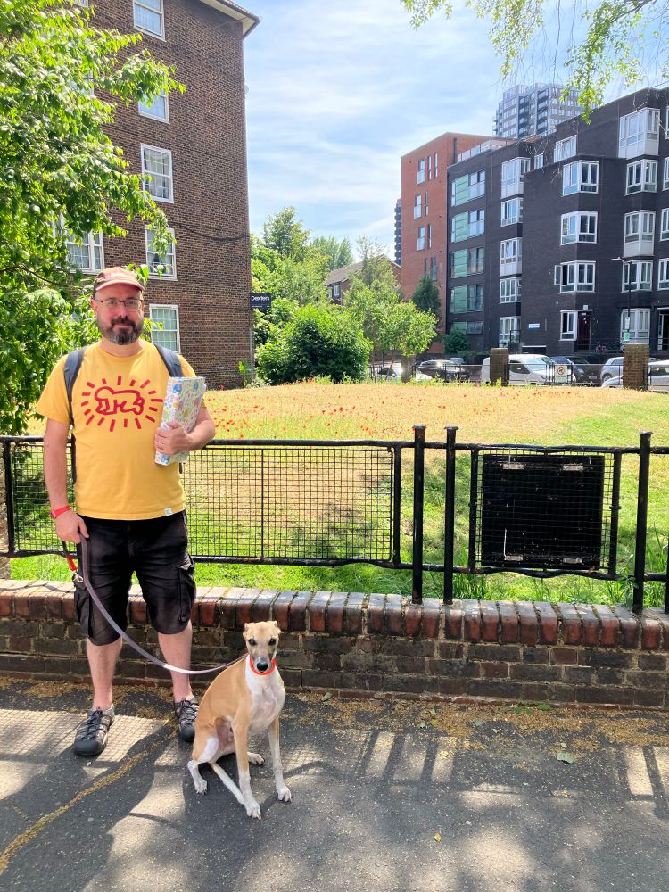 A bearded man in a hat with a yellow t-shirt on holds the lead of a seated dog. Behind them are railings made from Second World War stretchers and beyond is the outline of below ground bunkers.