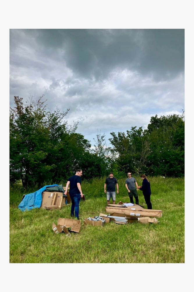 4 people stand around some boxes of kit looking for the instructions on how to build the tent structure.