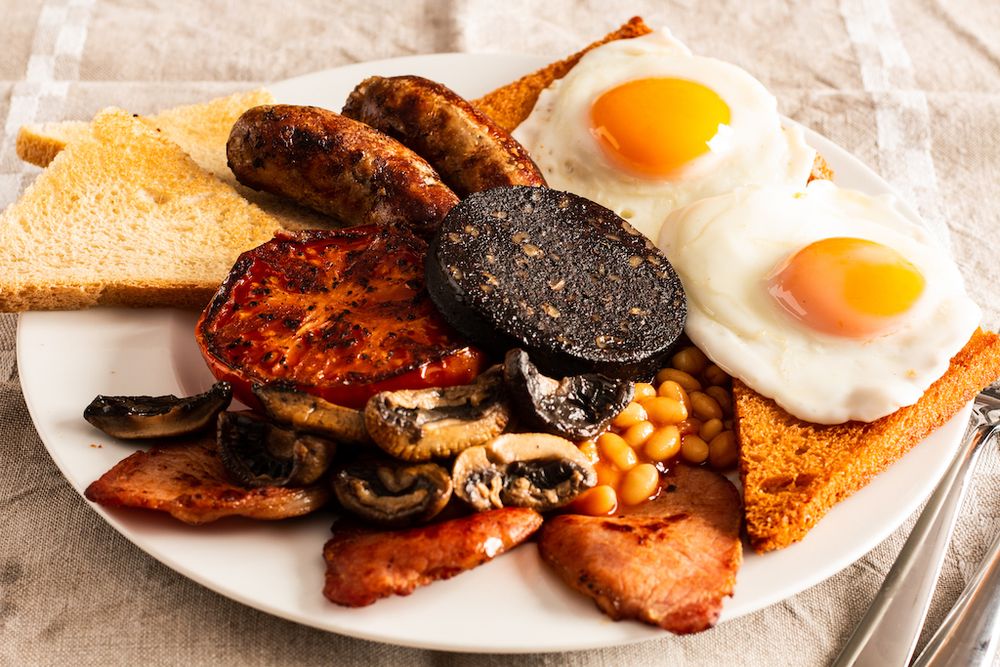 An image of a delicious Full English breakfast, including malt bread, black pudding, sausages, beans, tomatoes, mushrooms, baked beans and eggs sunny-side up