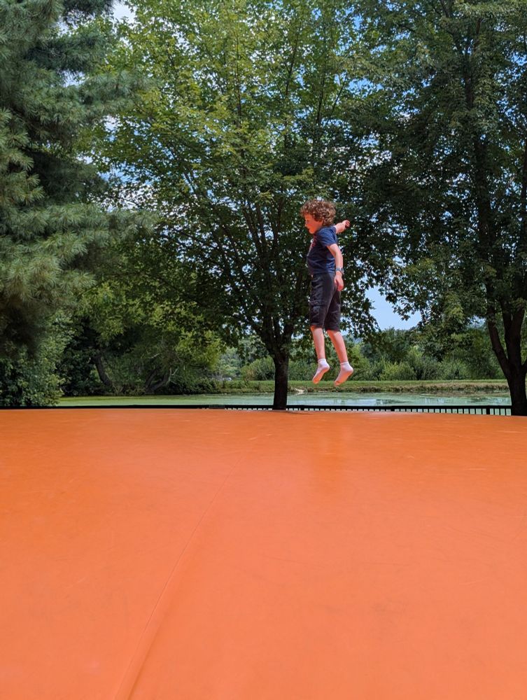 Young boy jumping on a giant outdoor pillow in front of a tree-lined pond