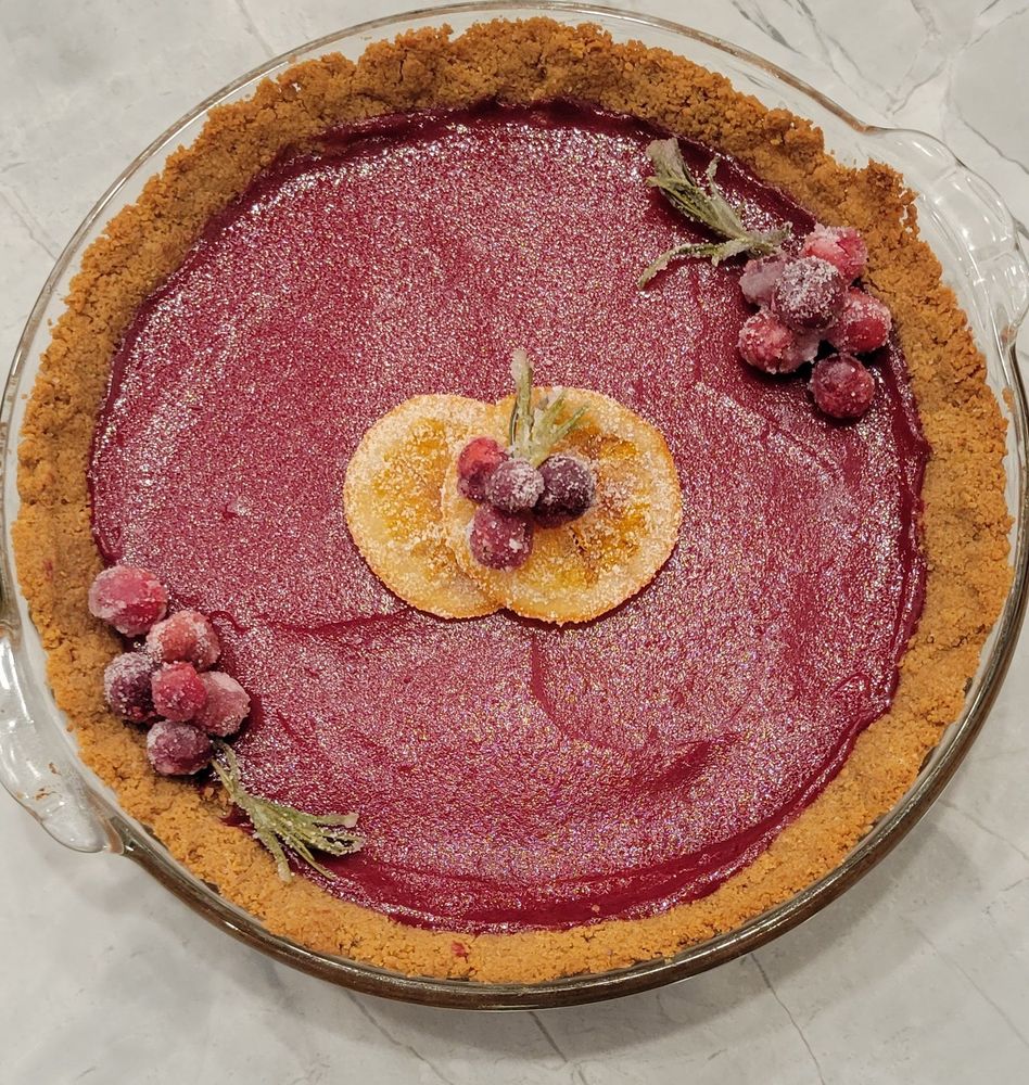 A cranberry curd tart with sugared rosemary and cranberry decorations as well as two candied orange slices in the center with more sugared berries.