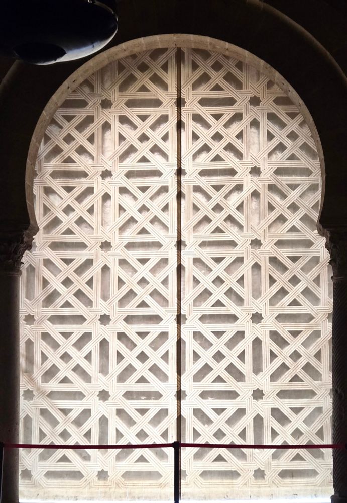 A keyhole shaped door, glowing white in the darkness. It has an 8 pointed star pattern where every pair of rays joins and crosses to form open x shapes before connecting to the next star.