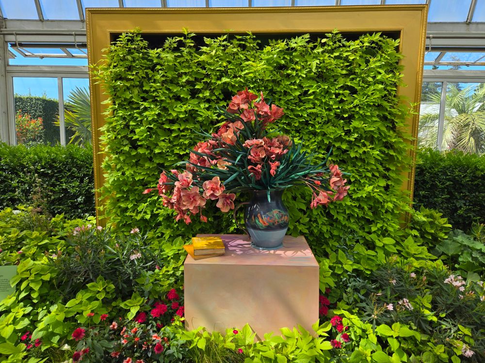 A metal sculpture of pink flowers in a vase next to two stacked yellow books. Meant to recreate a similar Van Gogh painting. The flowers sit before a field of leafy greens growing from a large gold frame.