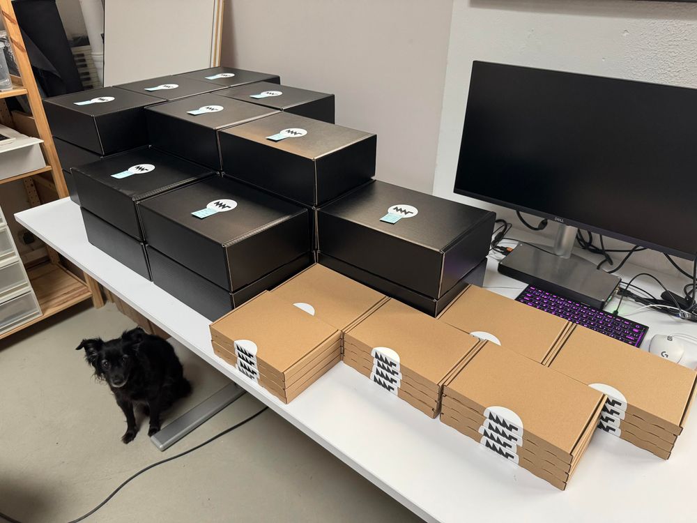 stacks of black boxes and cardboard boxes with MNT logos on them on a table at the studio. these contain MNT Pocket Reforms going soon to people who've ordered them in our shop. Tina the dog is lurking cutely under the table