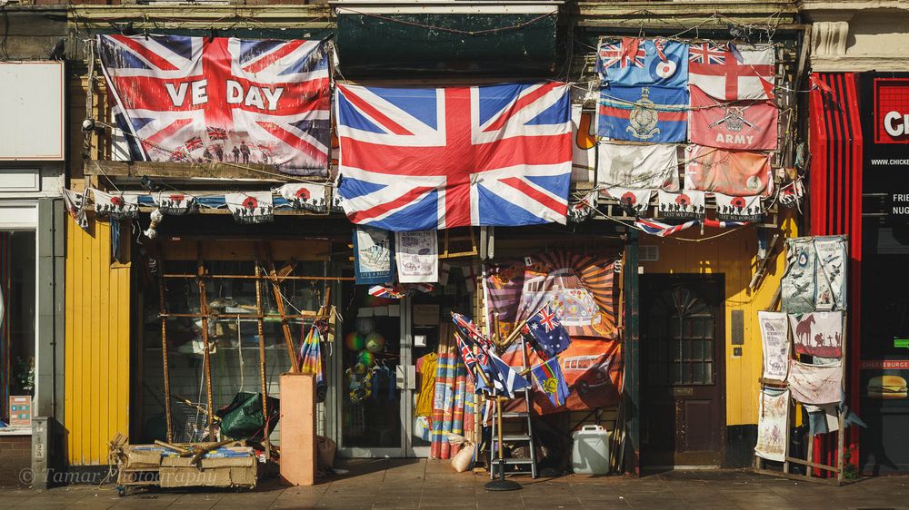 A colourful shopfront decorated with flags celebrating VE Day & various branches of the UK military.
