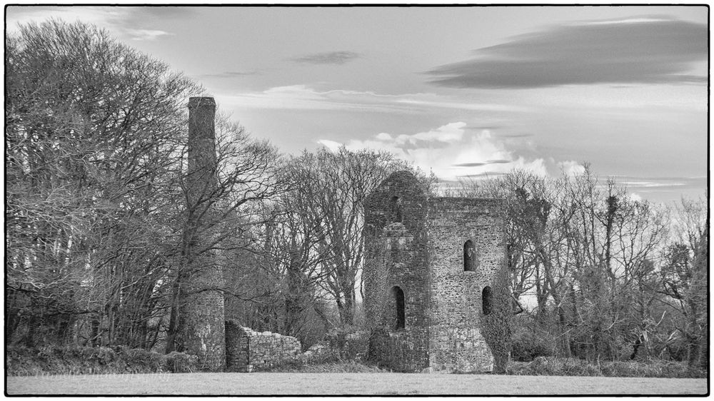 A monochrome photo of a long closed copper mine in Cornwall. In the centre can be seen the chimney for the boiler & to the right the old engine house. This would have housed the beam engine with a mine shaft just outside the building.