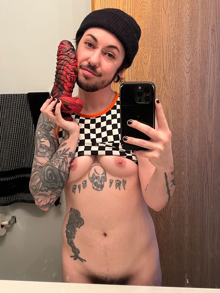 brick standing only wearing a tank top pulled over their tits. tattoos line their stomach. in one hand theyre holding the phone to take the mirror selfie. the other hand is holding an XL dildo to their face. theyre contentedly smiling. 