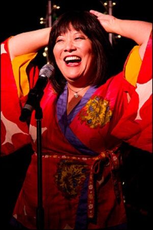 Ann Harada playing Christmas Eve in Avenue Q. Femme presenting actor of Asian descent in front of a mic wearing a red shirt/coat with purple trim and gold accents 