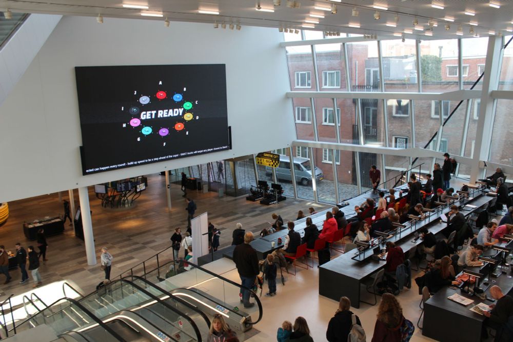 Ten people are sitting in front of large red arcade buttons in the main atrium of the Forum building in Groningen. In front of them is a large screen that has the lobby of Autogame Deluxe on. Around the players are more people standing and watching or visiting the Forum. All ten cars on the large screen are selected and in the center of the screen it says "Get Ready"