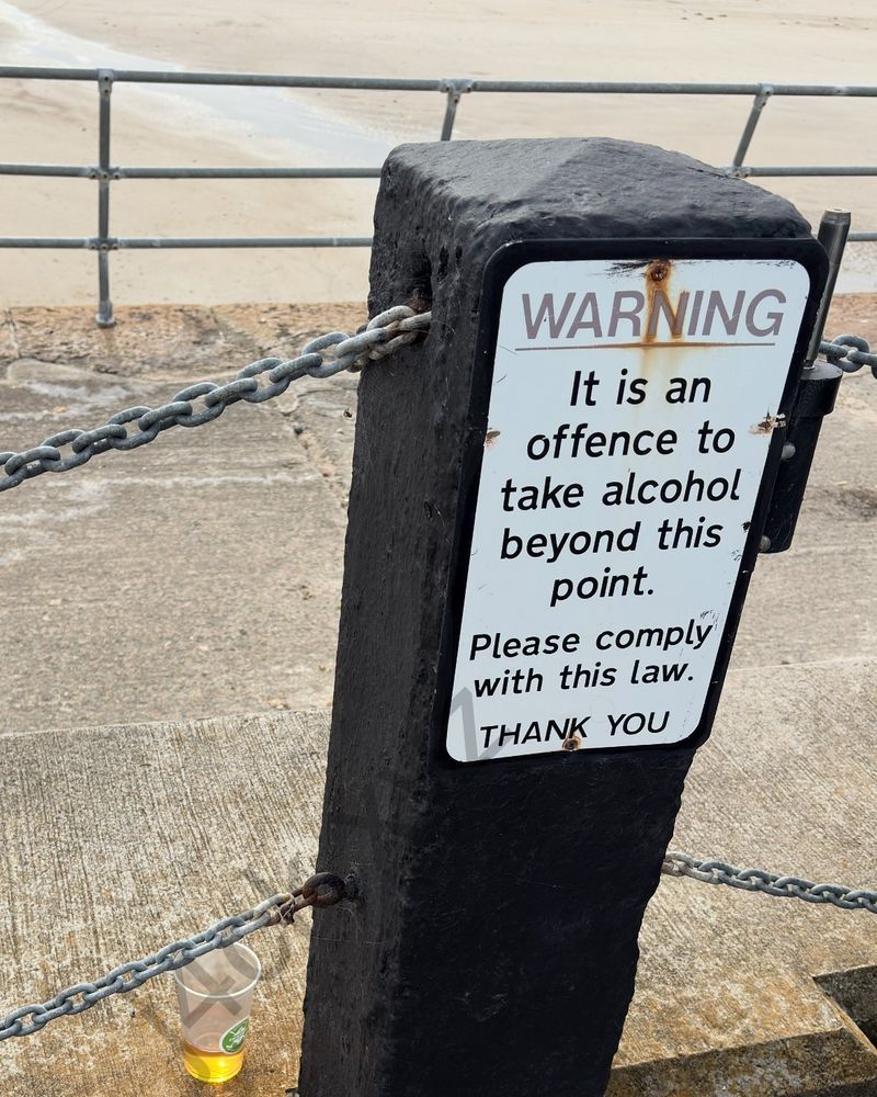 Sign “Warning: it is an offence to take alcohol beyond this point. Please comply with this law. THANK YOU” - on the left is a small cup of beer behind the chain of the sign.