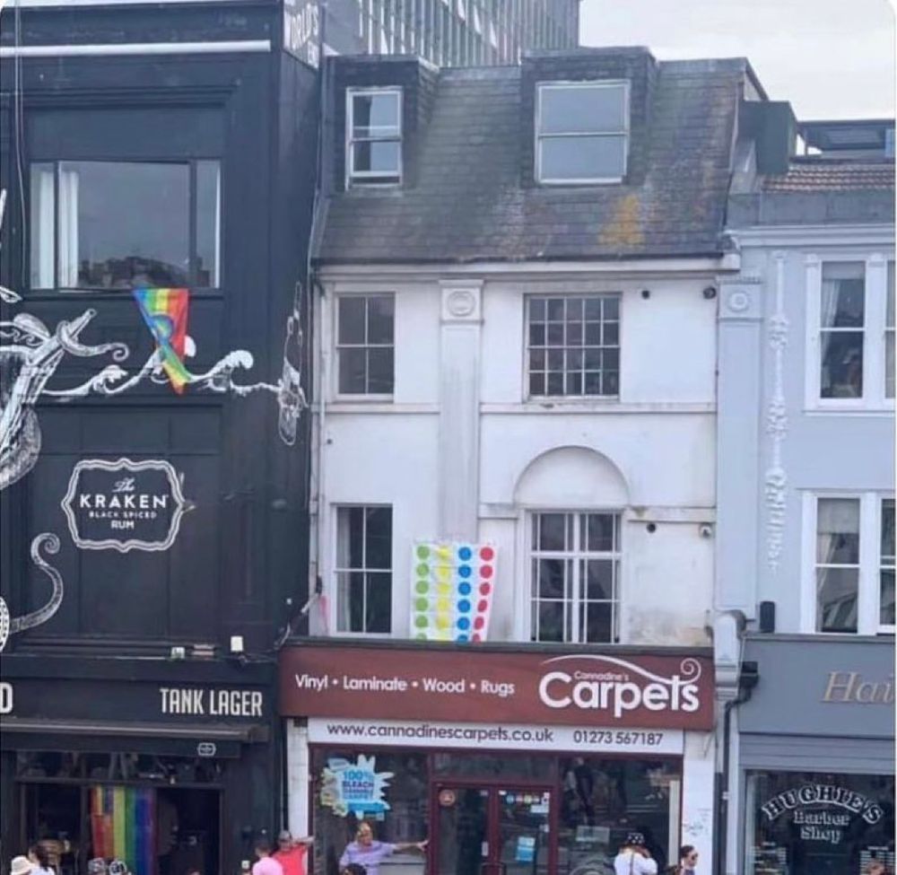 A multi-story building facade with a "Cannadine's Carpets" storefront, above which hangs a colourful twister-game mat. To the left, the building is painted black, with an advertisement for "The Kraken Black Spiced Rum." A pride flag is visible on the left.