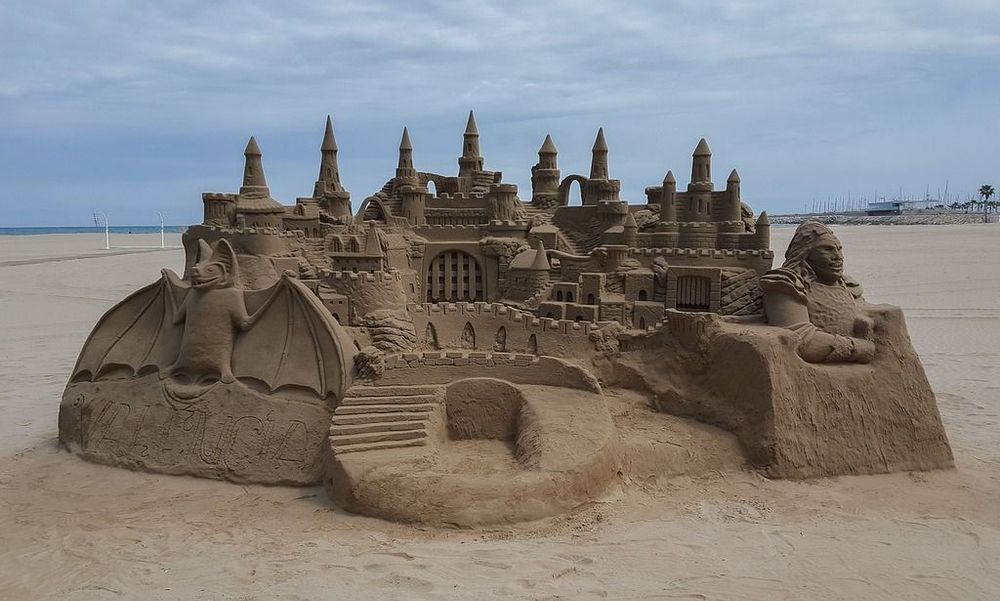 Photograph of a large sandcastle featuring multiple turrets, a sweeping staircase, and two elaborate figures flanking either side.