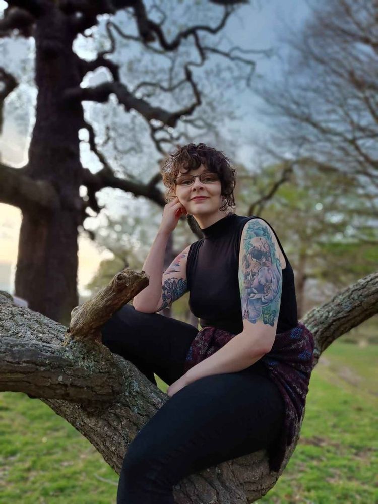 Jess, a person with curly brown hair and tattooed arms, sitting in a tree.