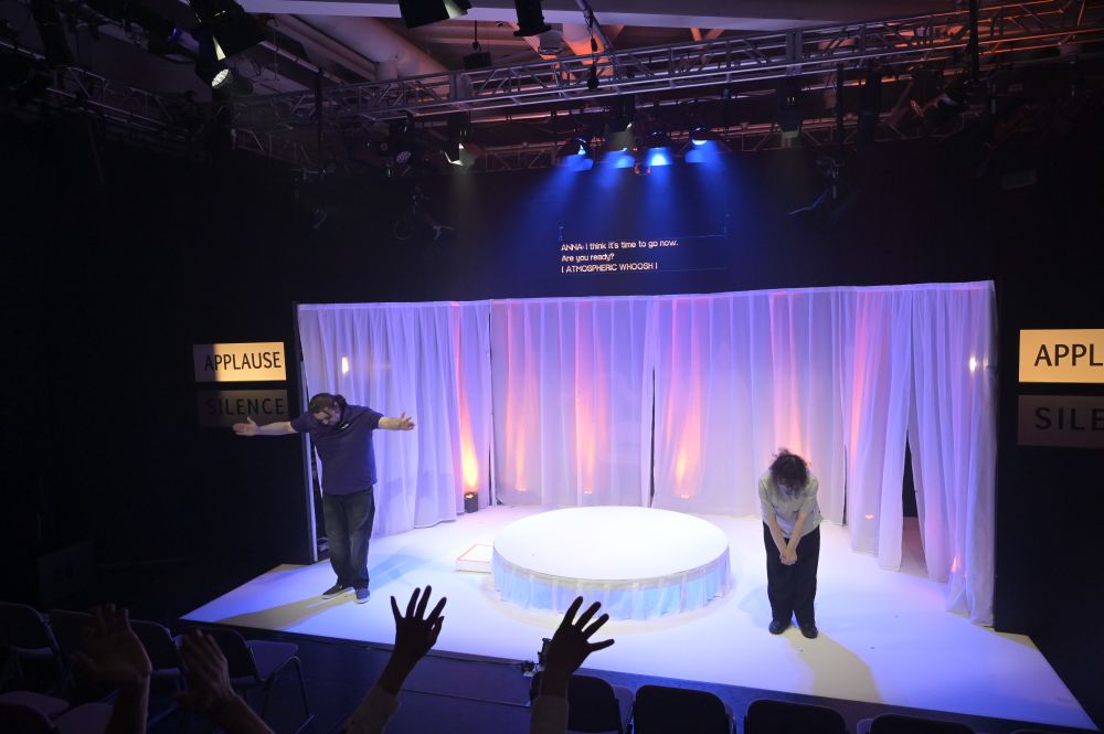 Two performers bow at the end of a stage performance. The stage is framed with white curtains and lit with soft blue and purple lights. A small round platform is centered on stage. Above, large captioned text reads: 'ANNA: I think it’s time to go now. Are you ready? [ATMOSPHERIC WHOOSH]'. Signs reading 'APPLAUSE' and 'SILENCE' are visible on both sides of the stage. Audience members' hands are raised in applause in the foreground.