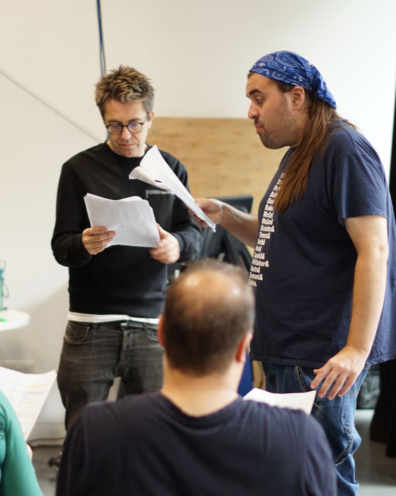 Nick, AAA Artistic Director, and Cian, AAA Associate Artist, are standing and discussing scripts in a rehearsal room. Nick on the left with short hair and glasses is reading his script. Cian on the right, with long hair and a blue bandana, is speaking animatedly while holding out his script. The back of a third person's head is visible in the foreground.