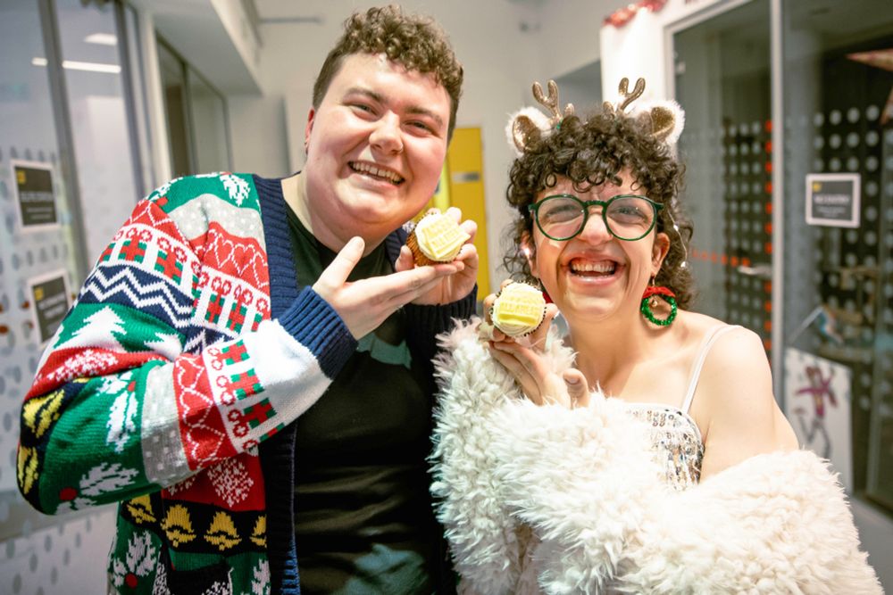 A cheerful, posed shot of a Diploma graduate and Anna, an Associate Artist, holding up cupcakes that read "ACCESS ALL AREAS." The graduate on the left wears a brightly colored Christmas cardigan. Anna on the right wears a fuzzy white coat, round glasses, and reindeer antlers, and is laughing.