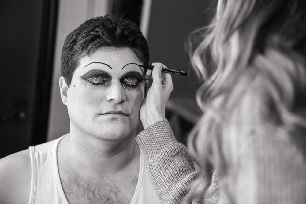 A close up black and white photograph of cast member Adam with their eyes closed with bold glitter eyeshadow and thin lined eyebrows being drawn on with a makeup pen.