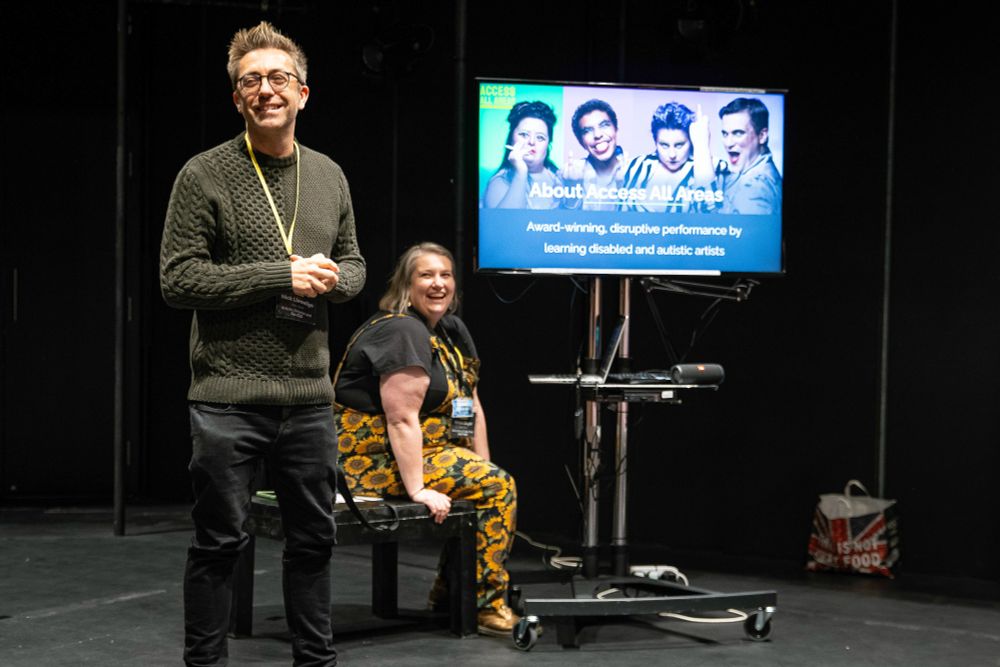 Nick and Helen lead a presentation in a dark studio space. Nick stands smiling in front of the audience while Helen  sits on a bench, also smiling. Behind them, a large screen displays photos of performers and the title “About Access All Areas: Award-winning, disruptive performance by learning disabled and autistic artists.”