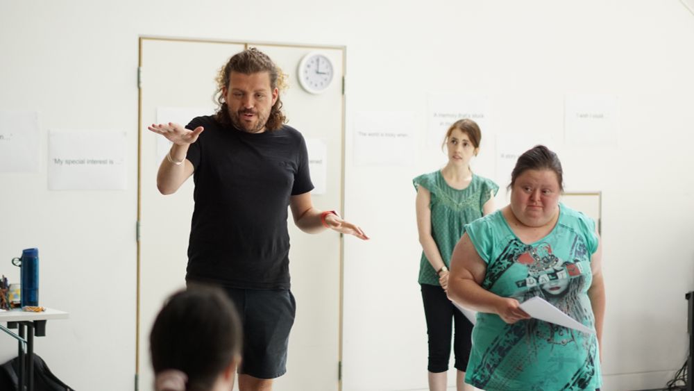 A group of people are engaged in a drama workshop in a bright, white-walled room. A man with curly hair and a beard, wearing a black T-shirt and shorts, gestures expressively while speaking to the group. A woman in a turquoise graphic T-shirt stands beside him holding a piece of paper, appearing thoughtful. Another woman in a green top and black trousers stands attentively in the background. Sheets of paper with printed prompts such as “My special interest is…” and “The world is tricky when…” are taped to the wall behind them.