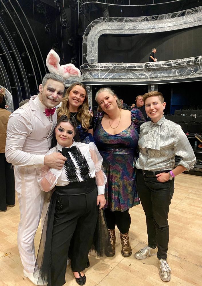 A Group of five people pose together on a theatre stage. Vito dressed as a white rabbit in makeup and bunny ears stands beside Ellie in a black and white costume with decorative black leaves. Three others stand beside them smiling — one in a shimmering multicoloured dress, another in a silver patterned shirt, and another in casual clothes. The stage behind them has silvery web-like decorations and theatre seating.