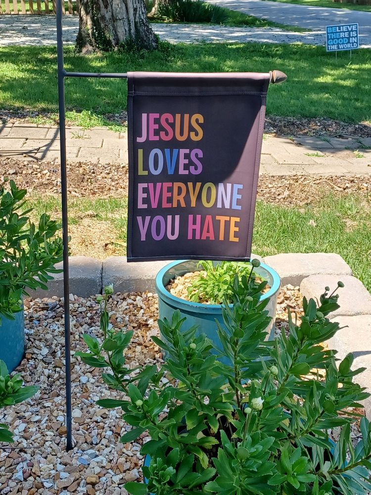 Black garden flag reads in colorful letters, "JESUS LOVES EVERYONE YOU HATE."