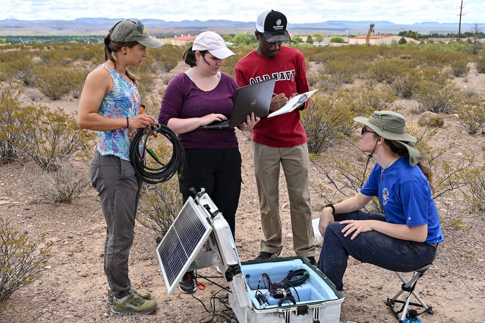 group of 4 interns working in a desert field covered or short shrubs with mountains off in the distance. intern on the left is is holding cables, while one in the middle holds a computer open and the other holds a clipboard while writing on it. The intern on the right is seated in a short stool with a metal box of equipment at their feet.