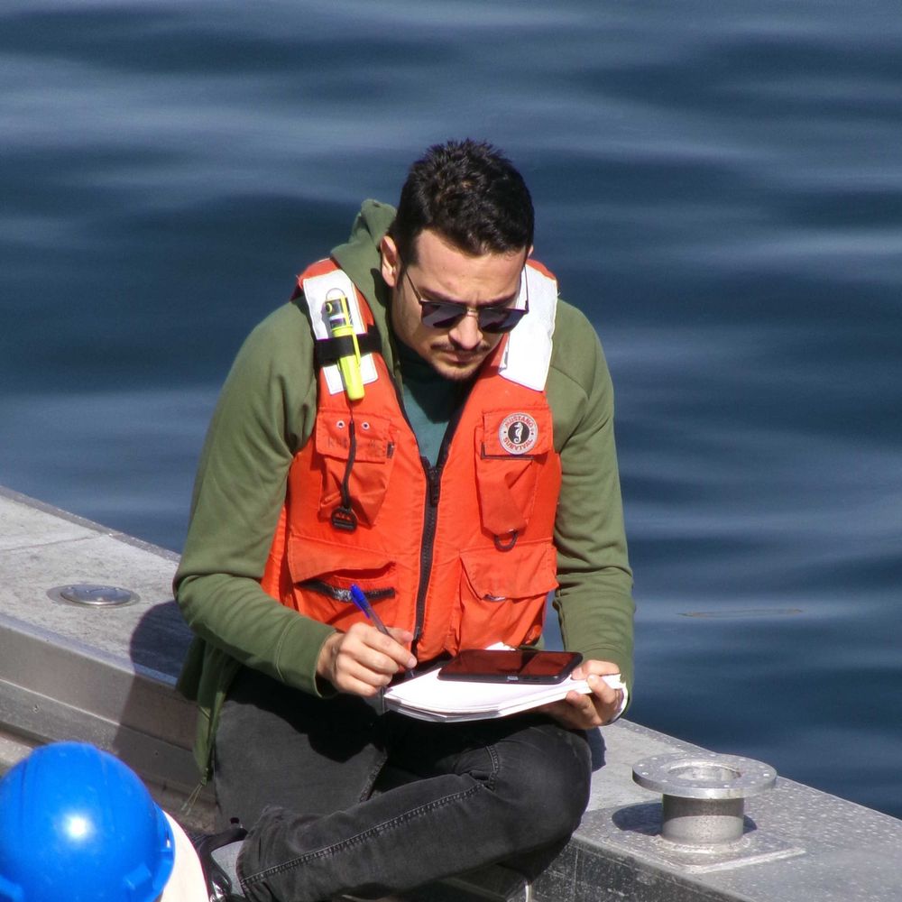 intern conducting field work at sea, sitting on the edge of a boat with a lifejacket on and the sea in the background. He is holding a pen in his right hand and papers in his lap, writing down information.
