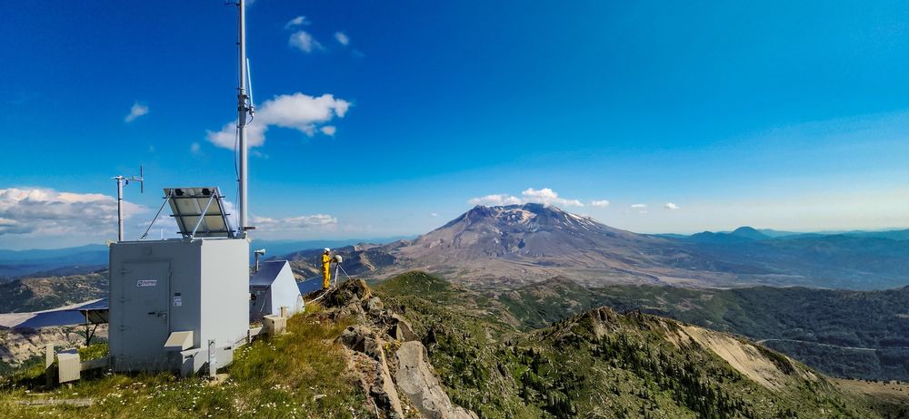 landscape photo of instrument and mount St Helen. On the left a large metal box with antenna and a solar panel sits, with a person in yellow working on an instrument to the right and a bit further back. To the right of that sits the volcano and its surrounding landscape. There is a bright blue sky above, with a few scattered clouds along the horizon and on the left side.