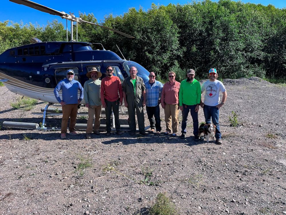 group of field workers stand together smiling with helicopter behind them. It sits on gravel and rock with bushy green trees filling the background and a small amount of clear blue sky peaking through the top. 