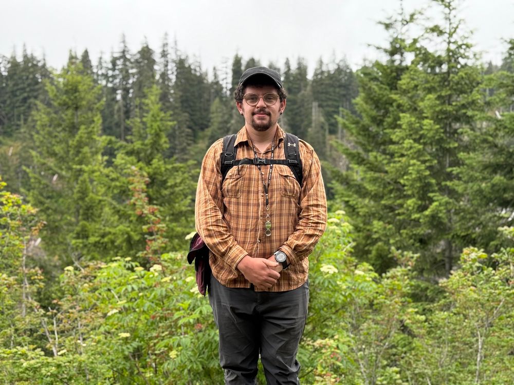 intern stands in middle of photo smiling in an orange plaid shirt with a backpack on. thick green foliage surrounds the background, with a hazy gray sky above.