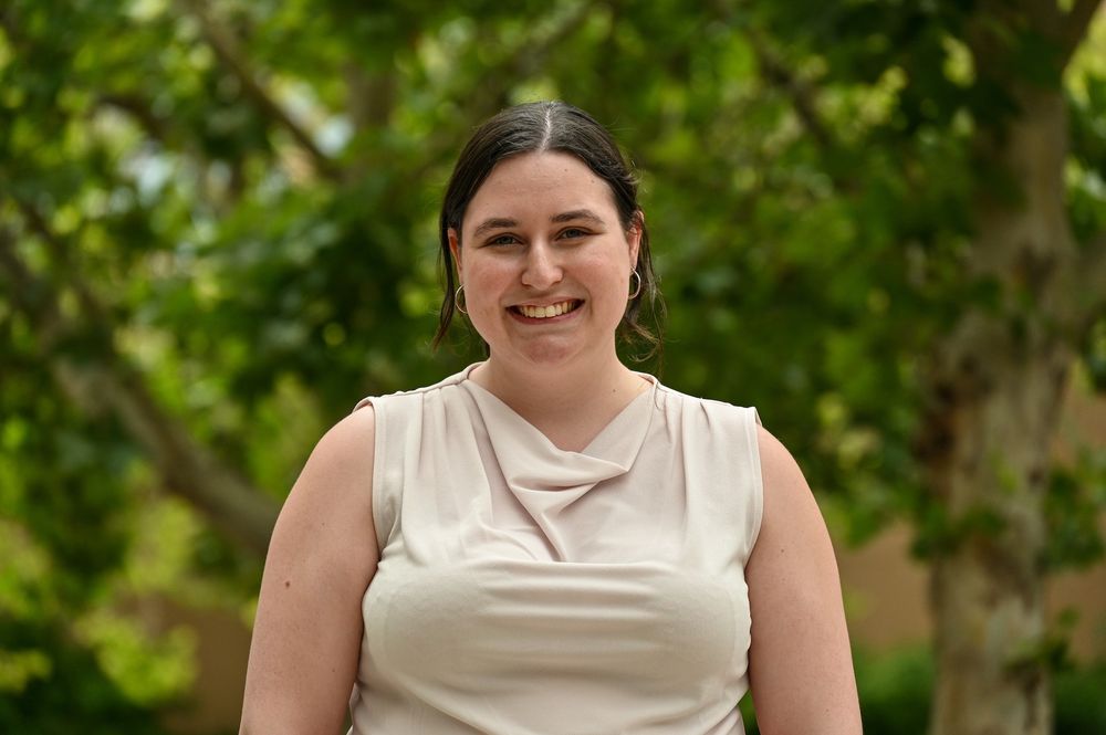 intern stands in center of frame facing the camera and smiling. behind her is green foliage.