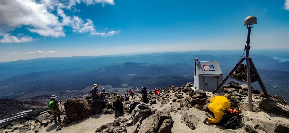landscape photo of the workers scatter around on rocks, and a man in yellow working on a large instrument on the right. The foreground consists of brown and gray gravel and rocks, with people scattered among them. the right side of the picture has a large metal gray box, and an instrument sitting on a tripod. A man in yellow in crouching down near the base of the instrument working on it. the background consists of rolling blue mountains, with a bright blue sky above and a cloud in the right side of the sky.
