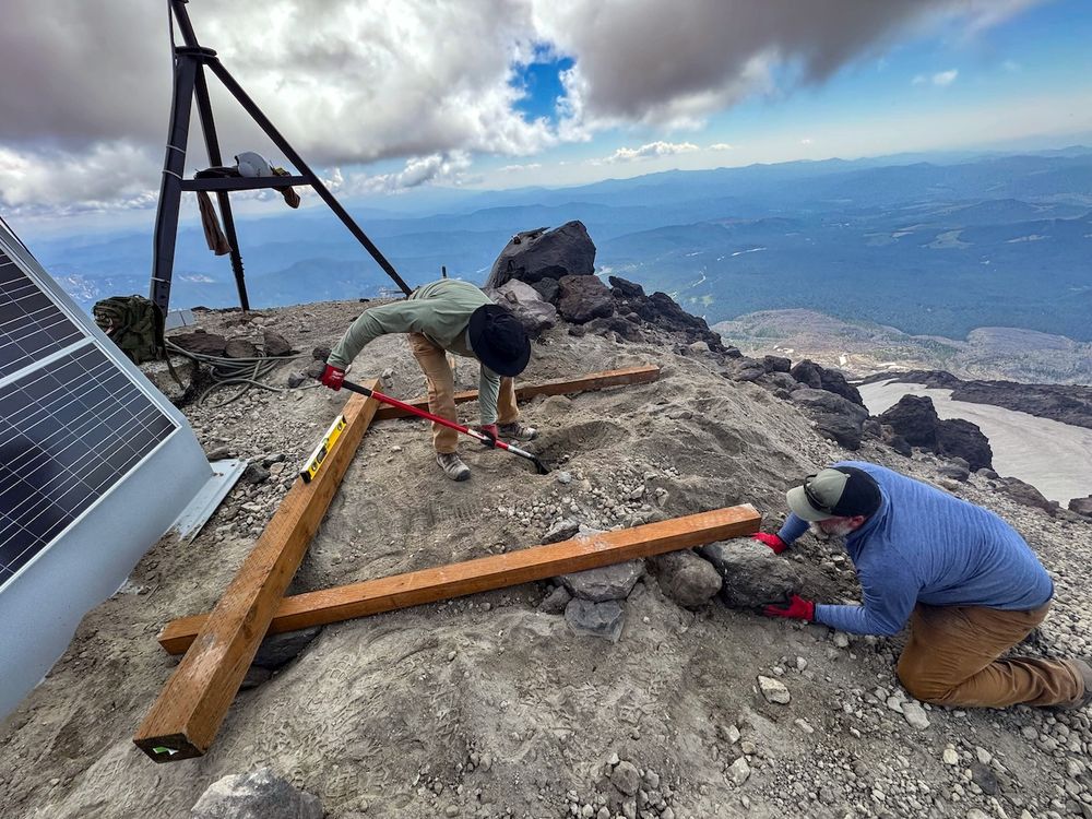 workers installing a platform for instruments on a brown and gray rocky surface. 3 wooden beams making 3 sides of a square are laying on the ground, with one worker placing rocks beneath a beam and the other digging a hole in the ground in the middle. to the left of them sits a large solar panel, and in the back left of the image sits an instrument on a tripod. in the background are rolling blue mountains, with large clouds filling most of the sky.