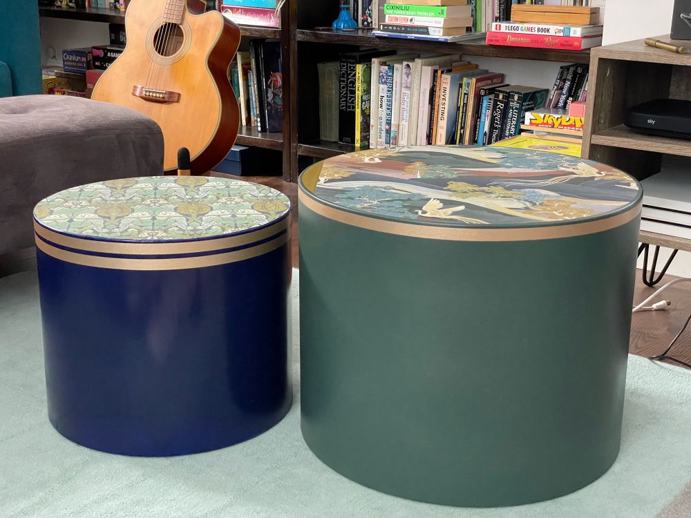Two cylindrical tables, one muted green, one blue, with patterned tops and gold banding close to the top edge. A guitar sits in the background looking tres artistique.