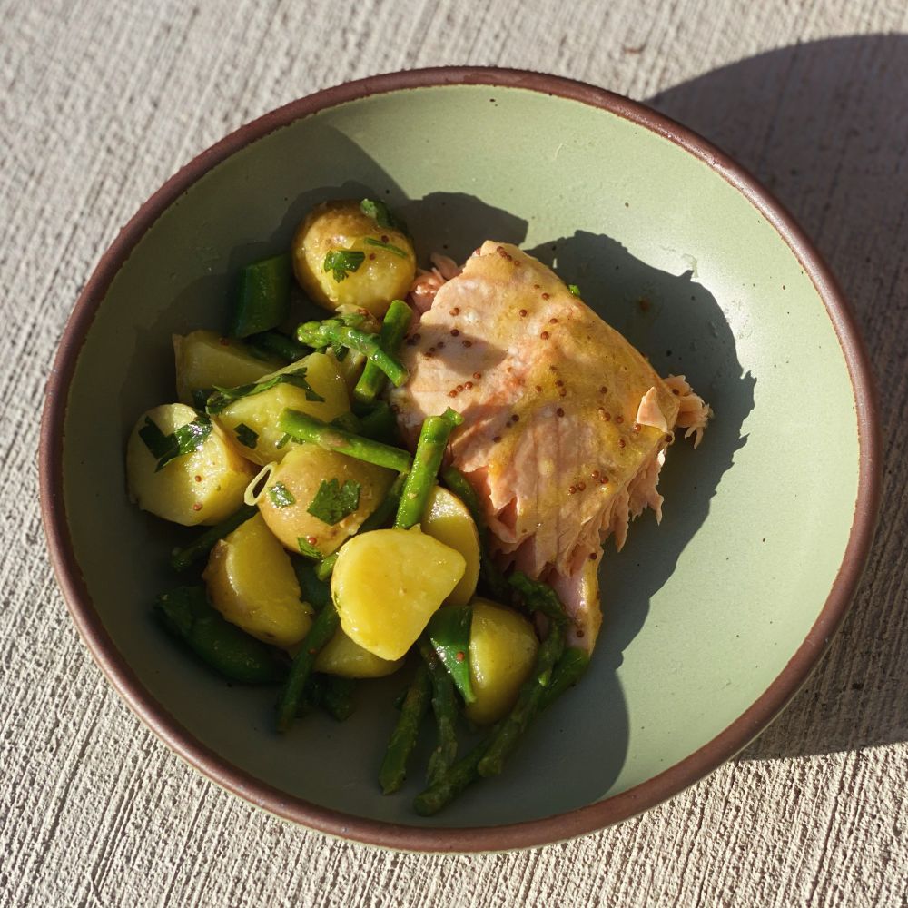 A pale green bowl with cooked salmon on the right side and potato salad with asparagus and herbs on the other side.