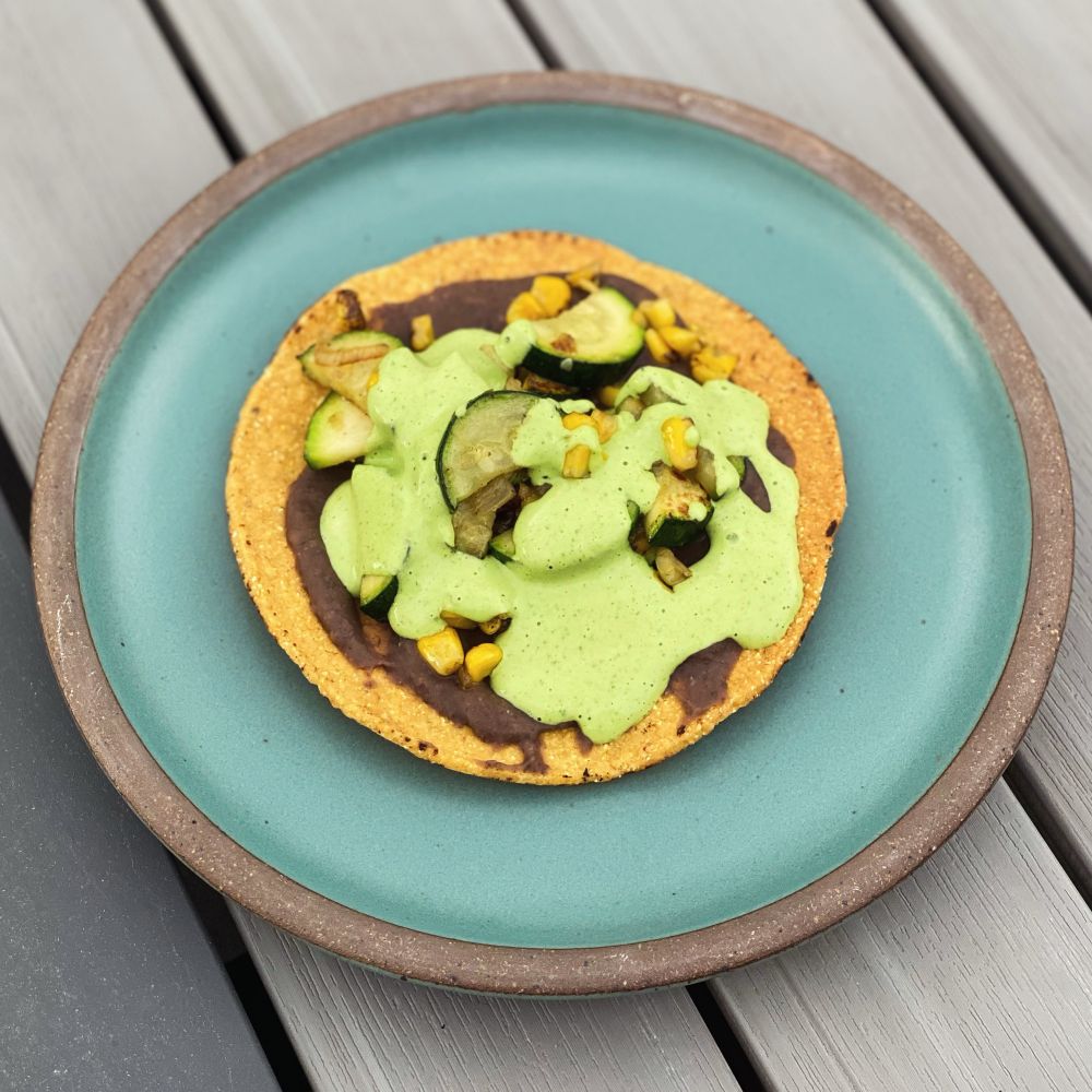 Tostada on an aqua plate. Layers of beans, veggies, and a bright green sauce.