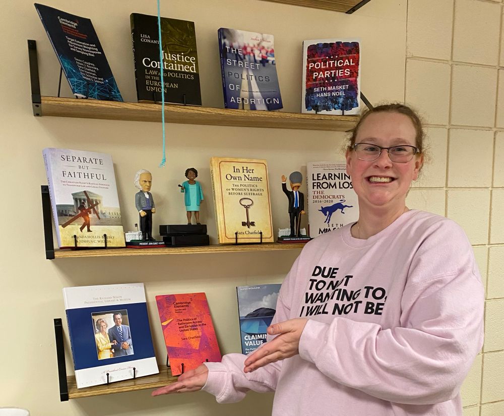 White woman in a pink sweatshirt stands Vanna White style in front of a set of shelves with books, indicating an orange/purple volume.