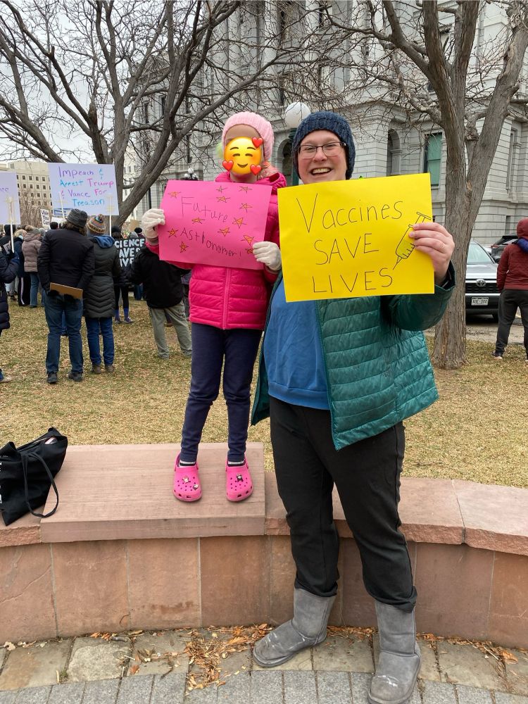 Woman in green coat holds sign reading “Vaccines Save Lives”. Next to her is a child with an emoji covering their face with a sign that reads “Future Astronomer!” They stand in front of a large white building, trees, and other people.